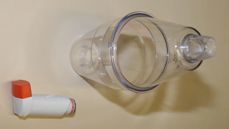 A large-volume plastic spacer device photographed next to a blue metered-dose salbutamol inhaler.