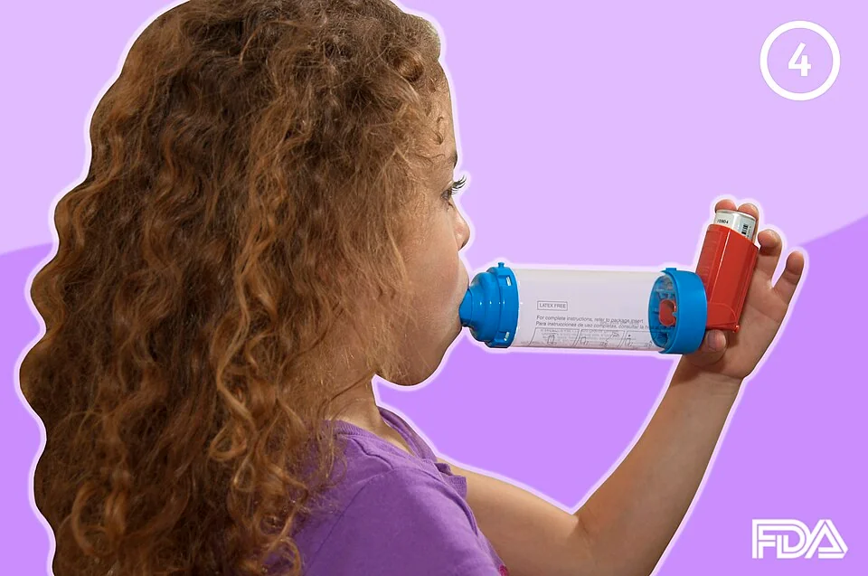 A child holding a pressurised metered-dose inhaler attached to a plastic spacer, with the mouthpiece of the spacer between her lips.