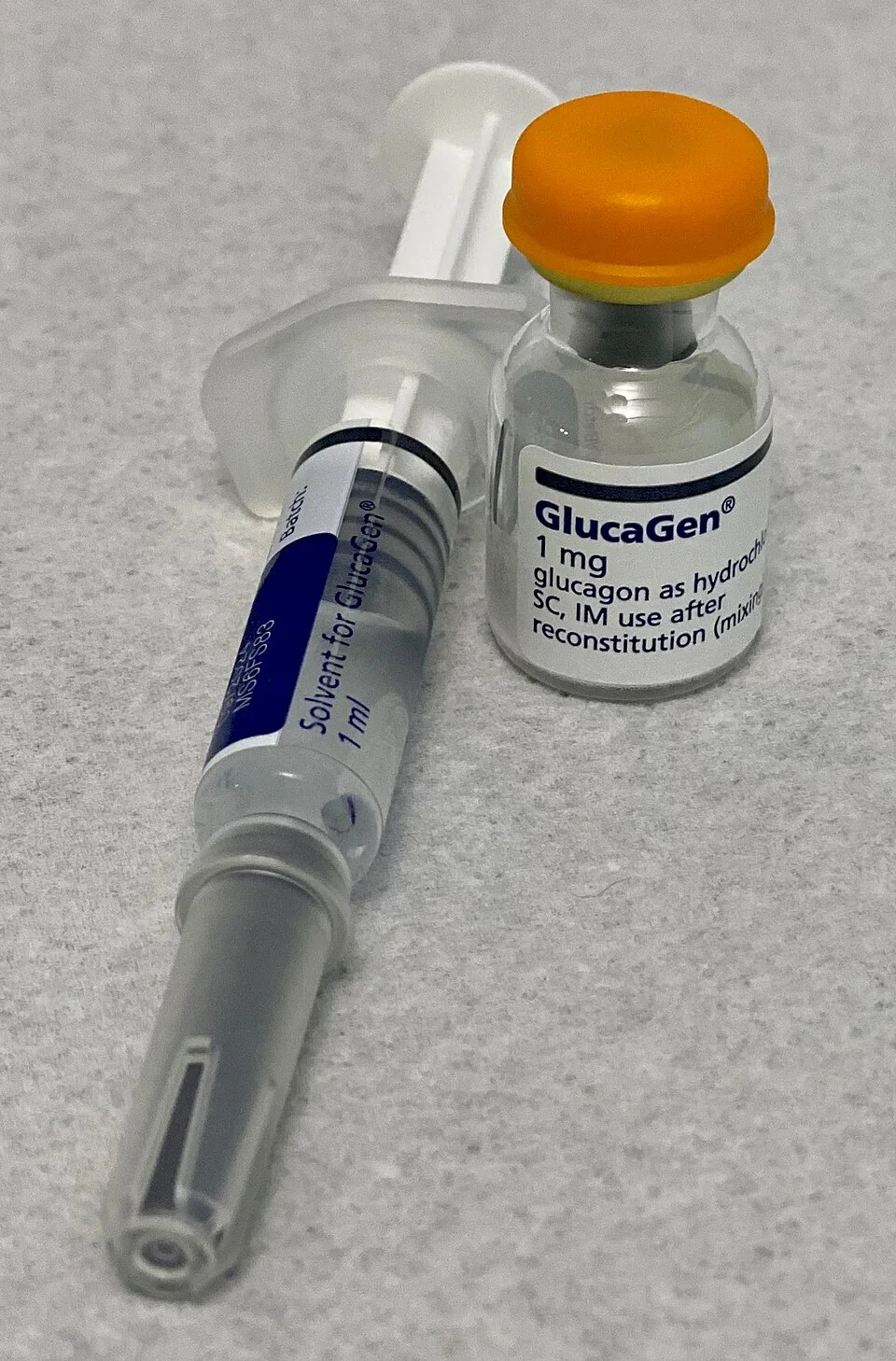 A glucagon injection kit photographed on a pale surface, showing a small glass vial of GlucaGen 1 mg powder with an orange cap alongside a prefilled syringe of 1 ml sterile solvent.