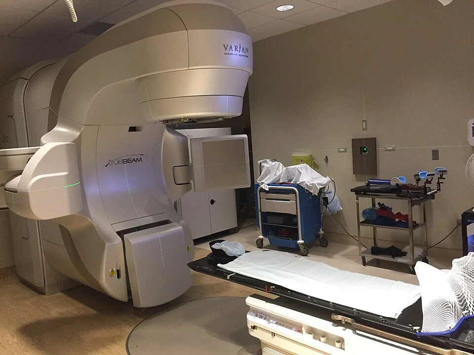 A medical linear accelerator in a treatment room, with a rotating gantry and treatment head positioned above a flat patient couch.